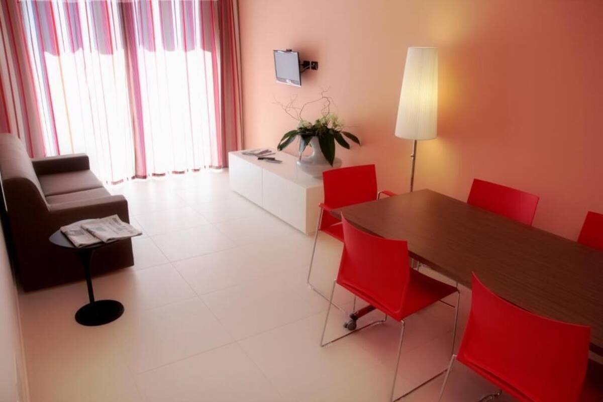 Uma sala de estar moderna com sofá, mesinha, mesa de jantar com cadeiras vermelhas, TV na parede e abajur.