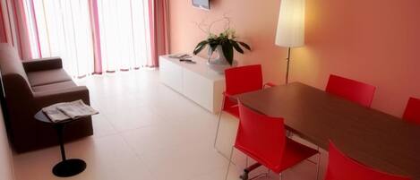A modern living room with a sofa, a small table, a dining table with red chairs, a TV mounted on the wall, and a lamp.