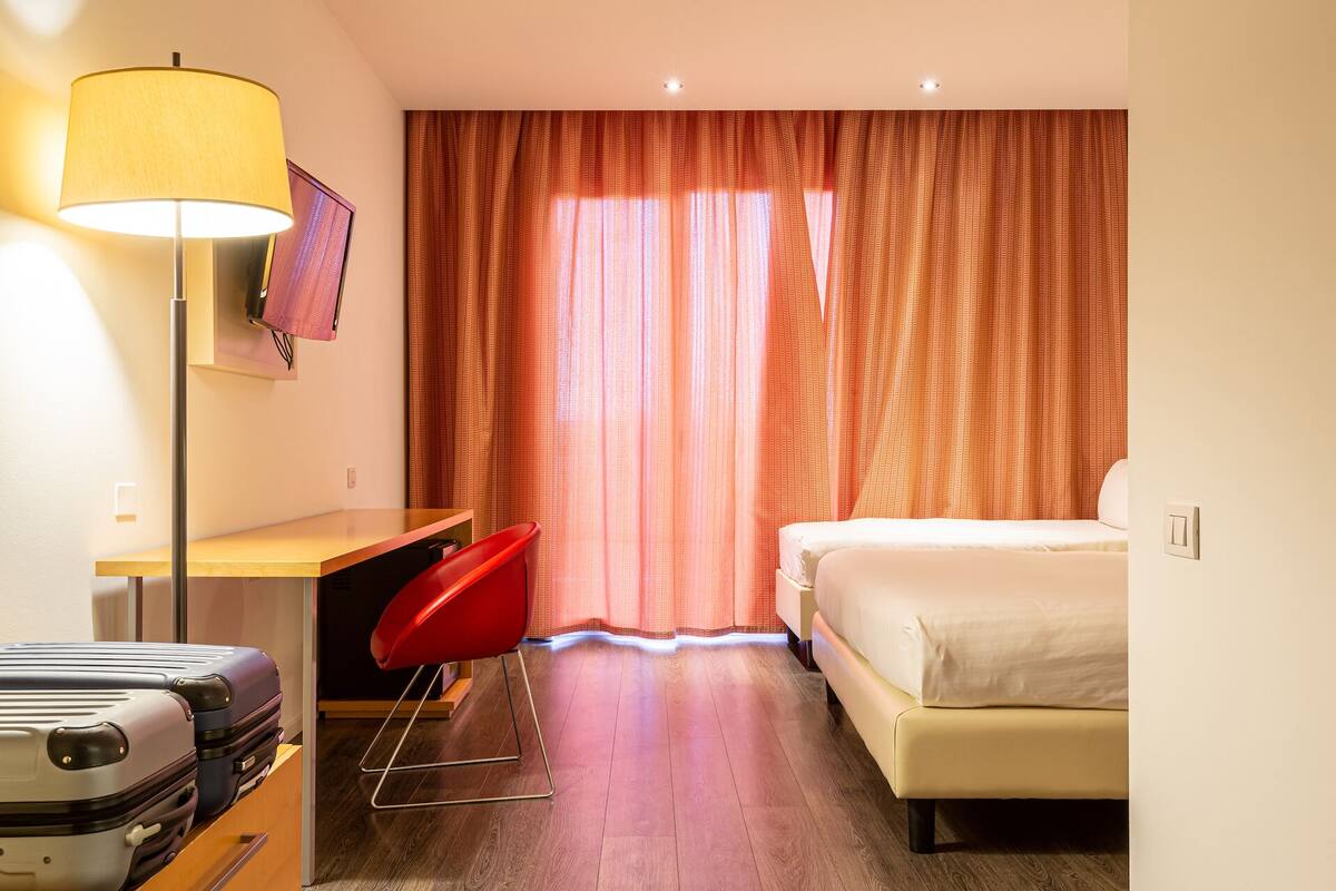 Quarto de hotel com cama, escrivaninha com cadeira, televisão e luminária.