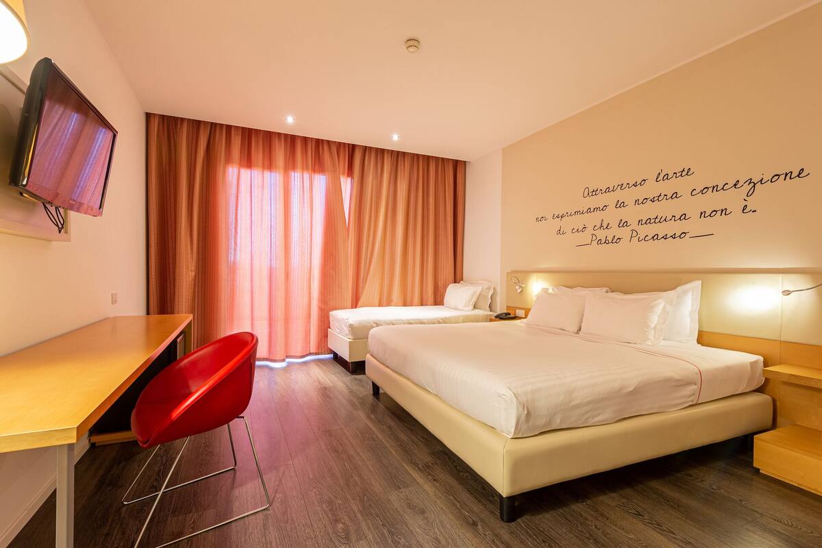 Quarto de hotel com duas camas, uma escrivaninha, uma cadeira vermelha e uma parede com uma citação.