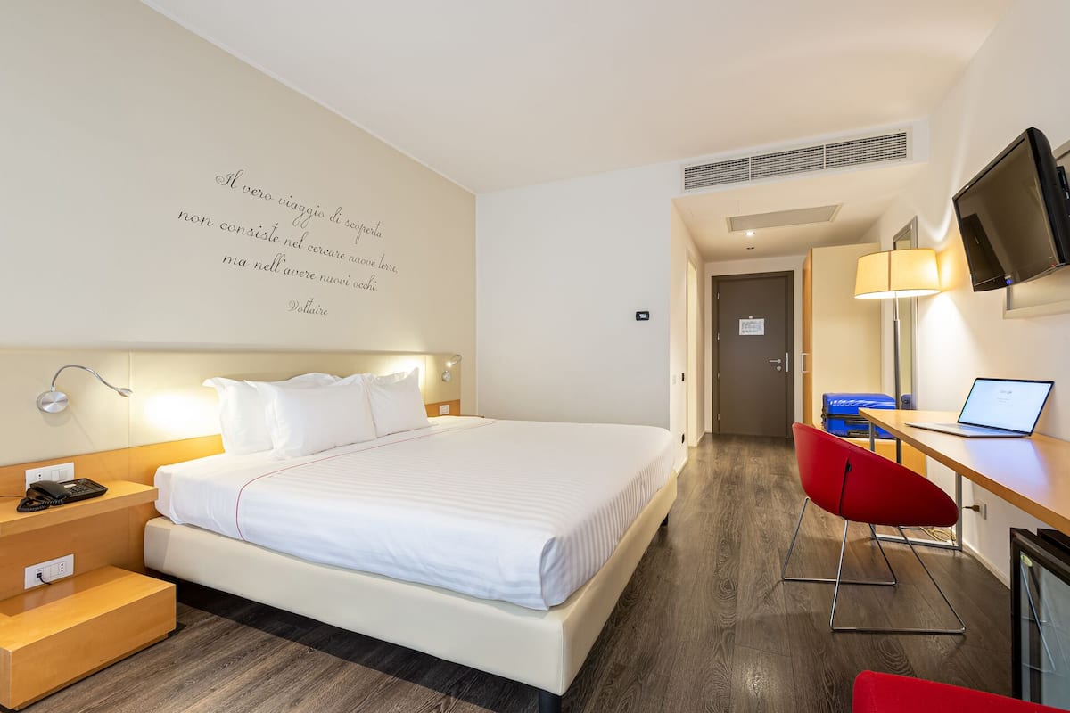 Quarto de hotel com uma cama grande, uma escrivaninha com um laptop, uma televisão e uma cadeira vermelha.