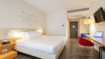 A hotel room with a bed, bedside table, lamp, TV, desk, and chair.