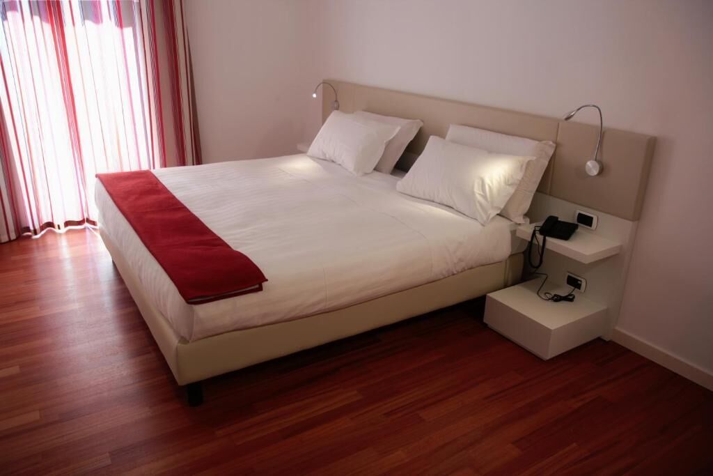 A neatly made bed with a red throw, a bedside table with a phone and charger, and a nightstand with a lamp.