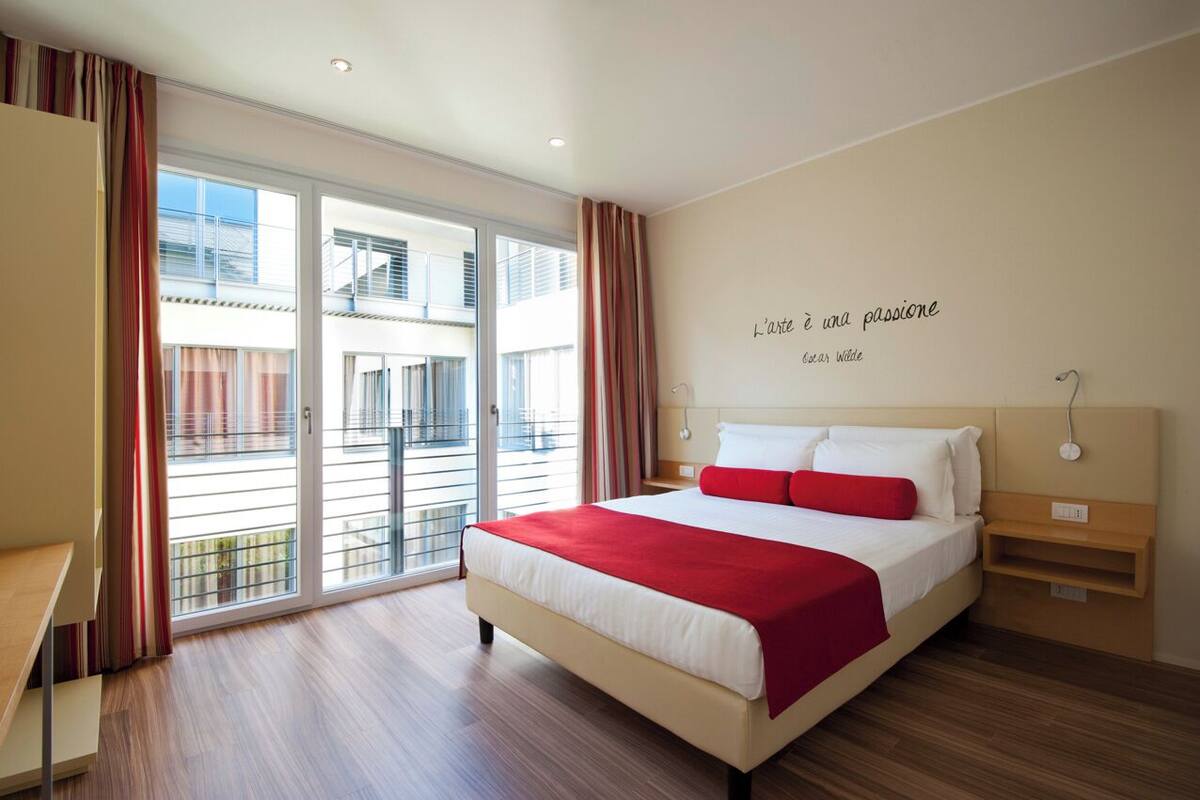 Quarto de hotel moderno com uma cama grande, uma escrivaninha e vista da cidade pelas portas da varanda.