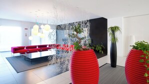 Un espace d’accueil moderne avec un coin salon rouge, un mur décoratif agrémenté d’éléments suspendus et de grandes plantes en pot.