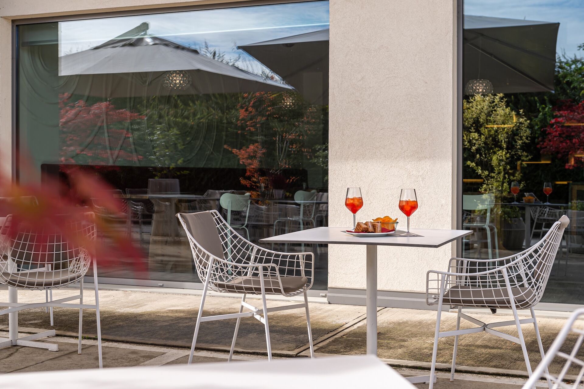 An outdoor seating area with two white wire chairs, a table, and two glasses of red drink.