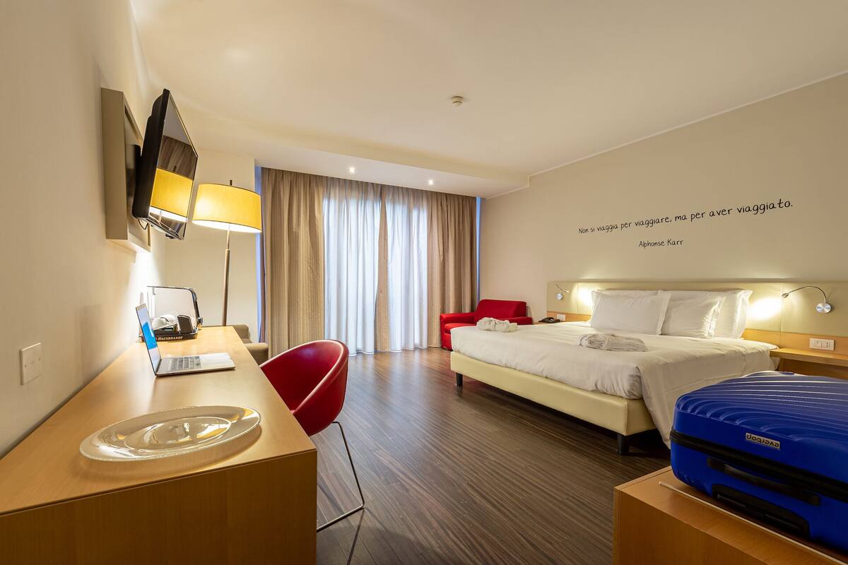 Quarto de hotel com cama, escrivaninha, cadeira e mala.
