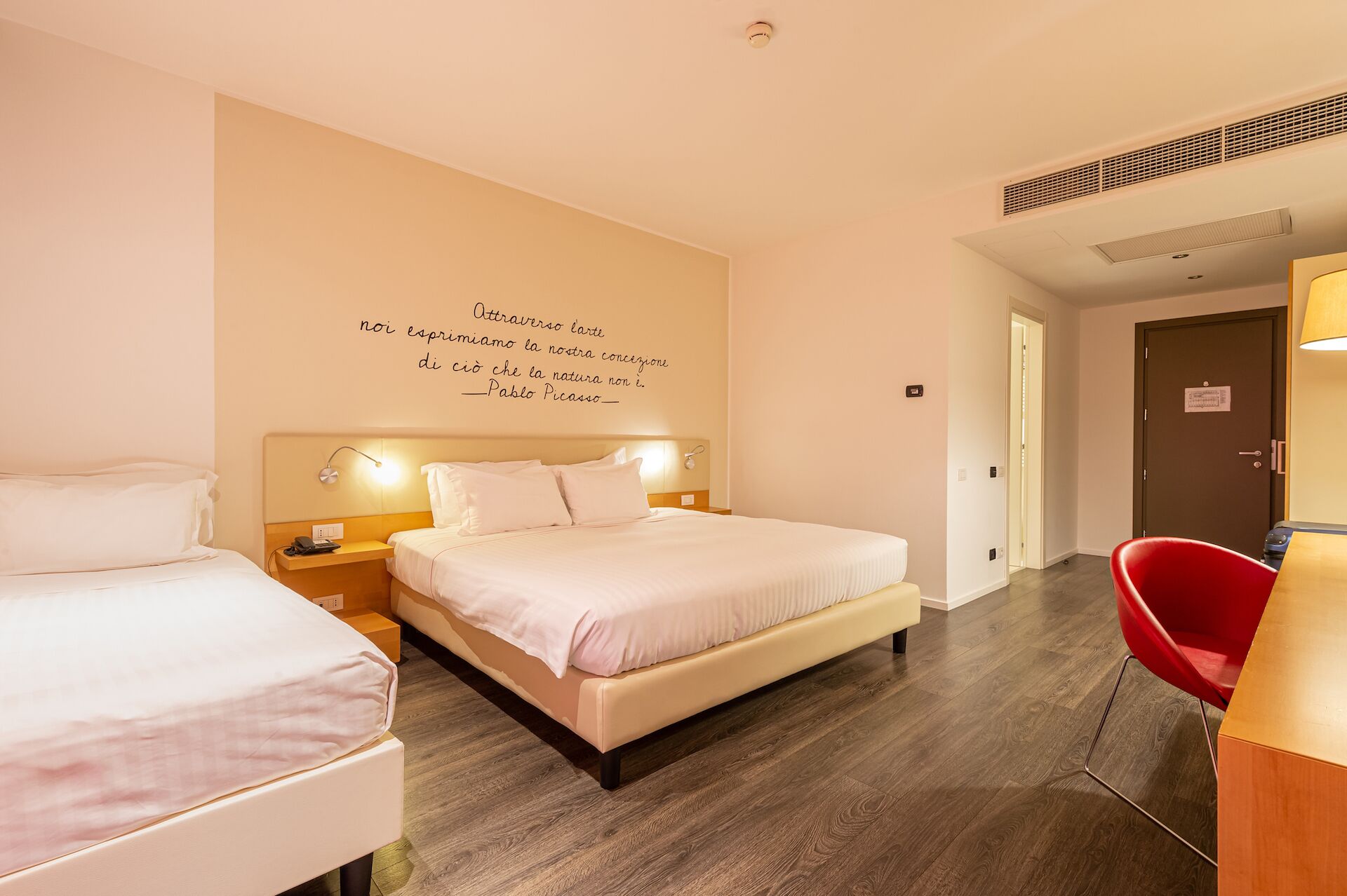 A hotel room with a bed, bedside tables, a red chair, and a wall with a quote.