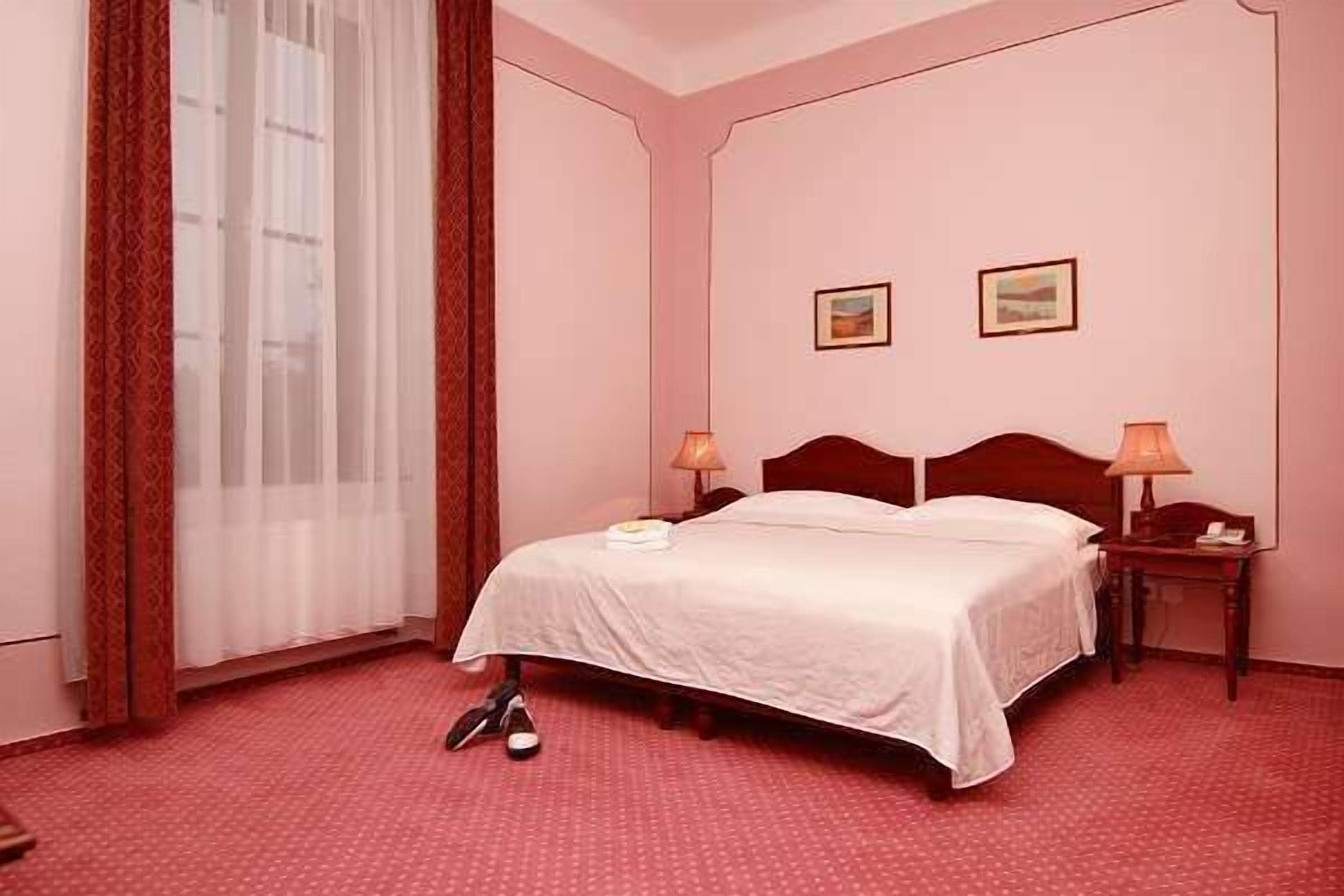Une chambre d’hôtel avec un lit, deux tables de chevet, une fenêtre avec des rideaux légers et un sol recouvert d’une moquette rouge.