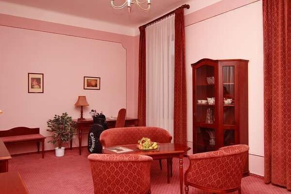 A room with a red carpet, a wooden cabinet, two chairs, a table, a lamp, and two framed pictures on the wall. - Golf Resort Hotel Konopiste (Bystrice)