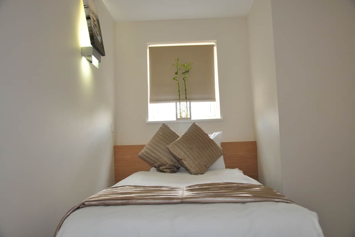 A neatly made bed with a headboard, a window with blinds, and a wall-mounted light fixture.