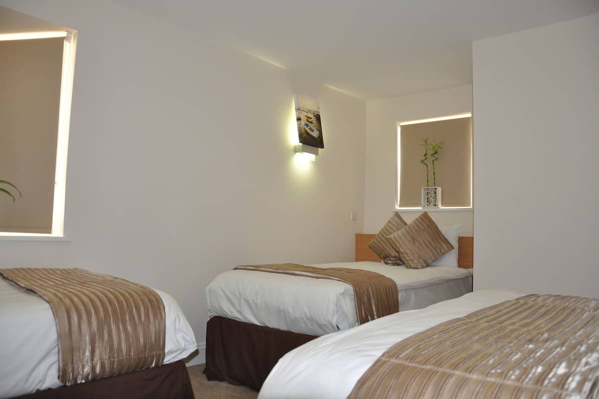 A hotel room with three beds, each with a striped blanket, a wall-mounted light, and a mirror reflecting a plant.