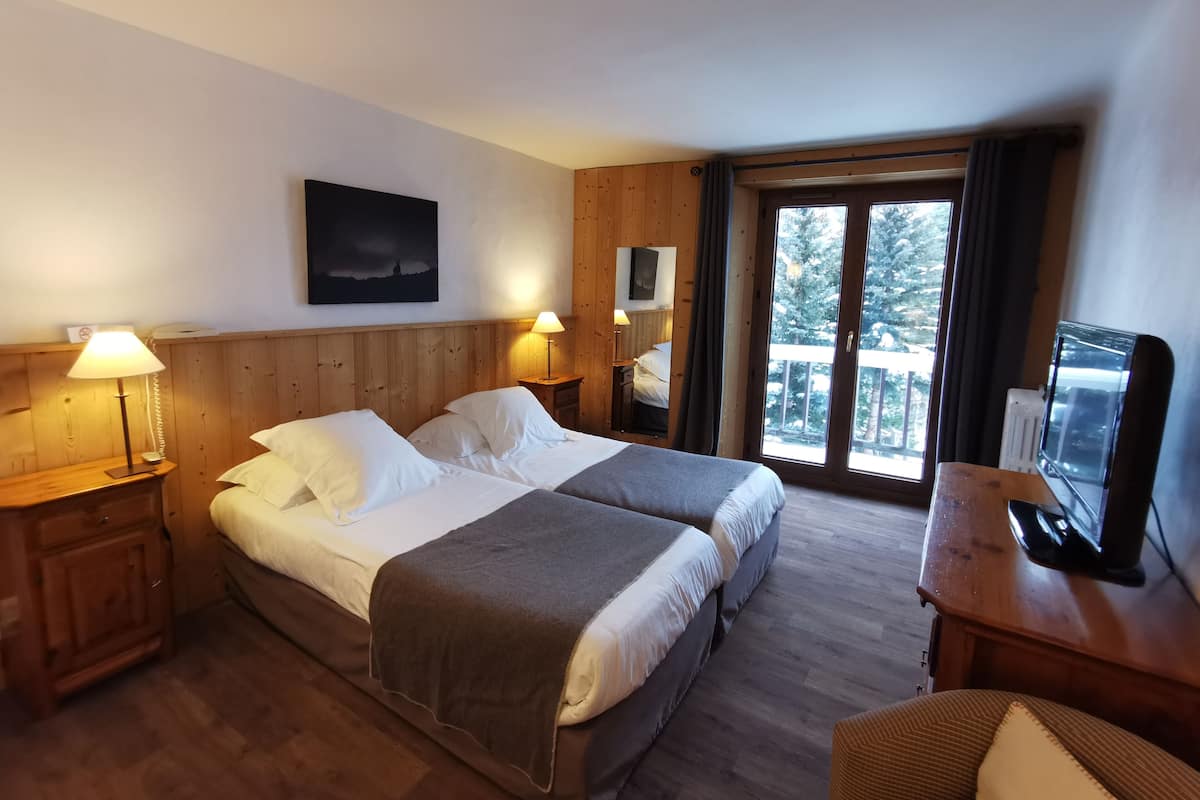 A hotel room with two beds, wooden headboards, a TV, and a balcony with a view of trees.