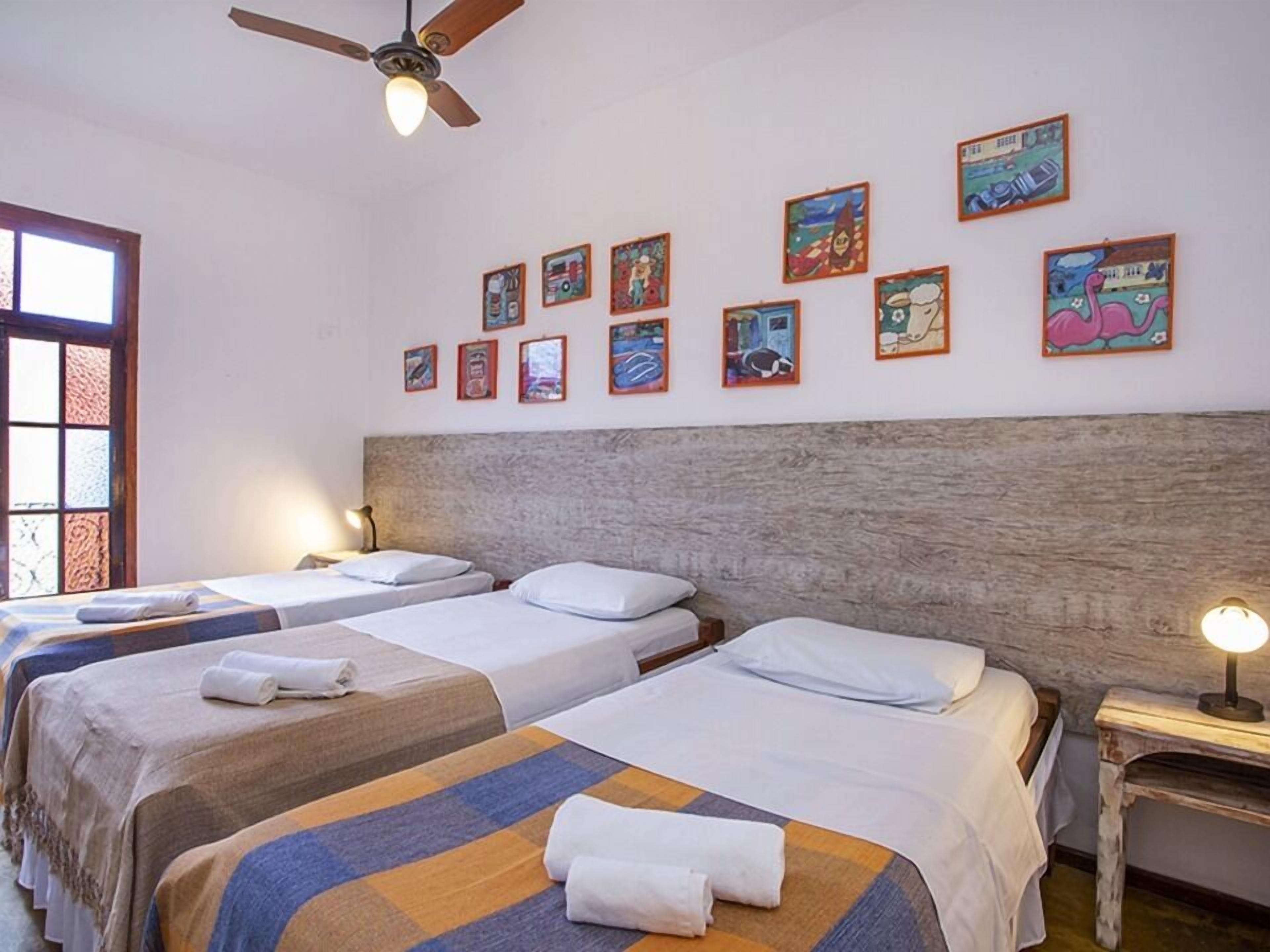 Une chambre d’hôtel avec trois lits simples, une tête de lit en bois et un ventilateur de plafond.