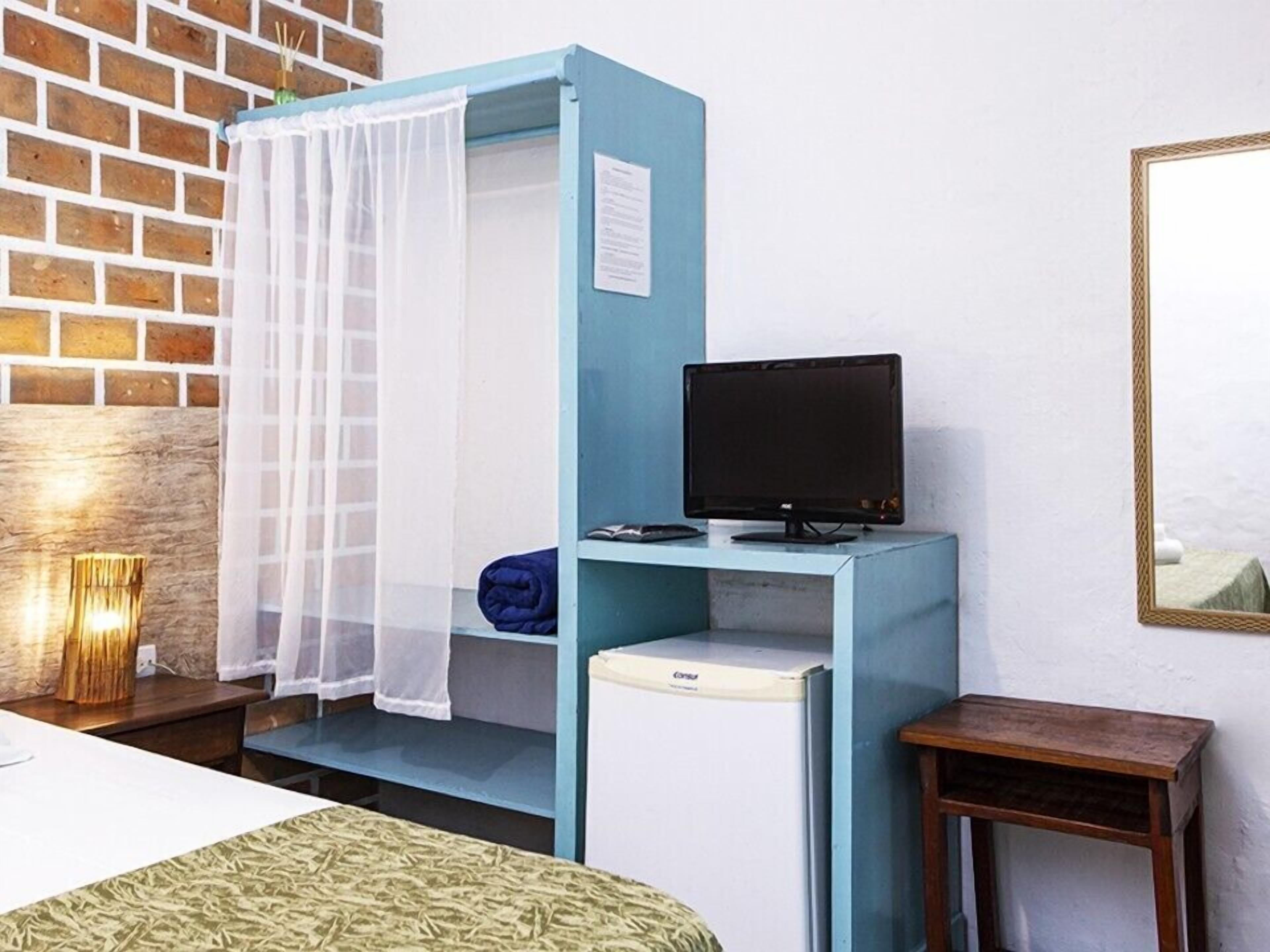 Una habitación con una cama, un armario azul con televisor, una mesita y una pared de ladrillo.