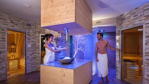 A spa area with wooden walls, a central wooden structure with a bowl, and two people in towels. - Hotel La Torretta (Vermiglio)
