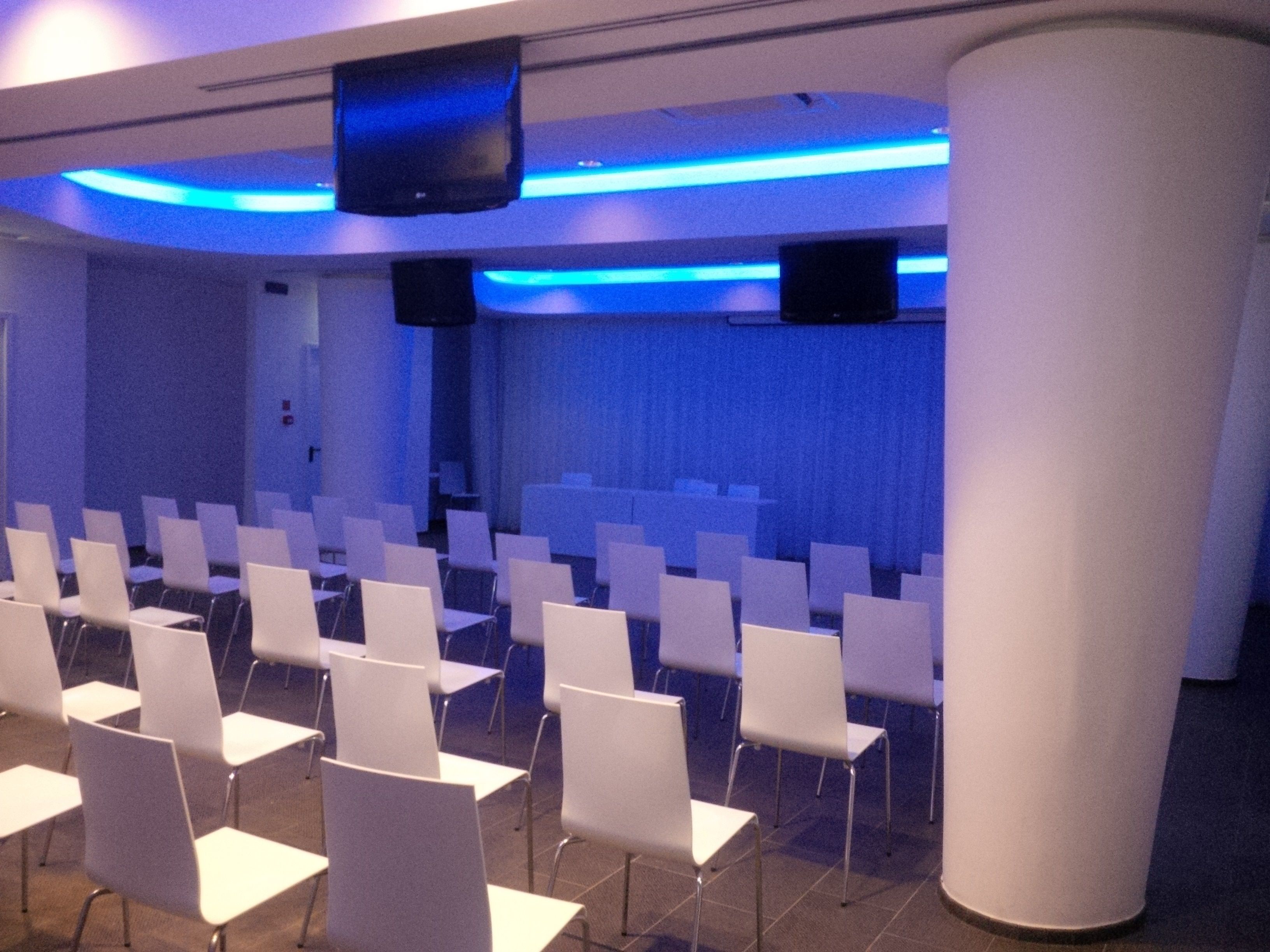 A conference room with rows of white chairs, a large screen, and blue ambient lighting.