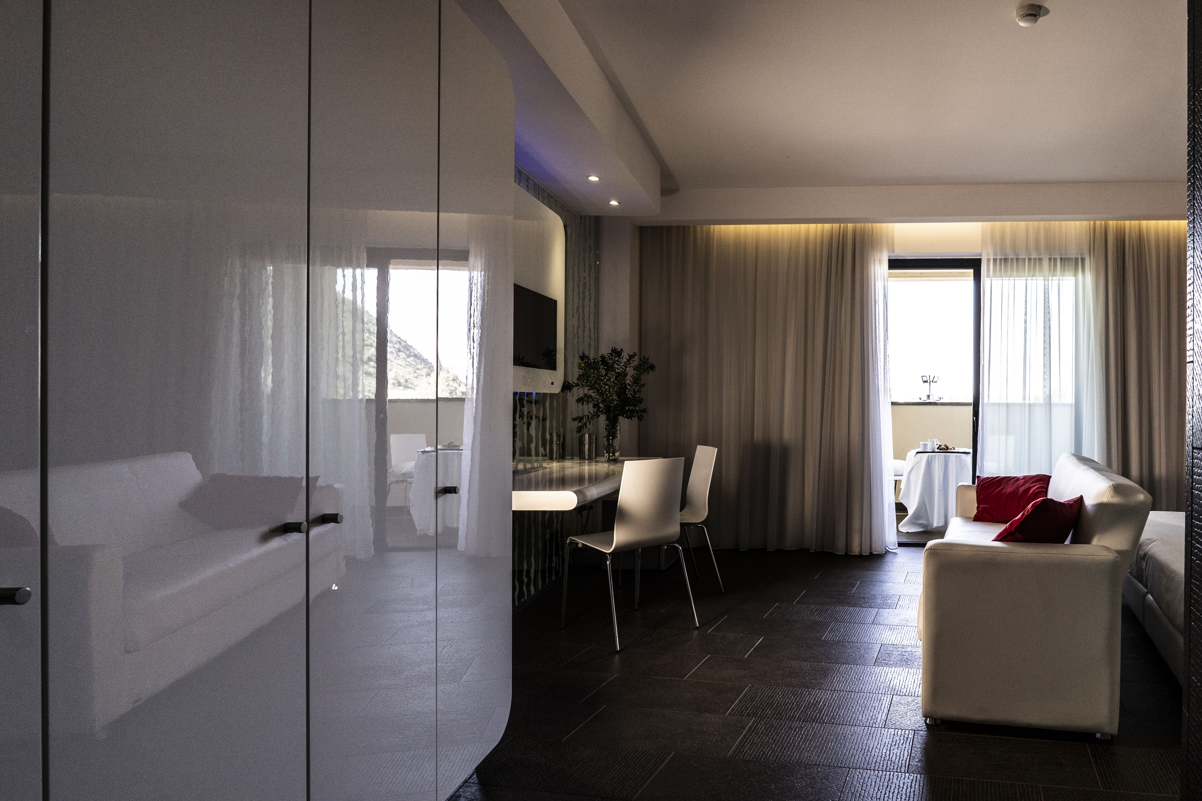 Una habitación de hotel moderna con un separador de vidrio, un sofá, una mesa de comedor con sillas y un ventanal con cortinas.