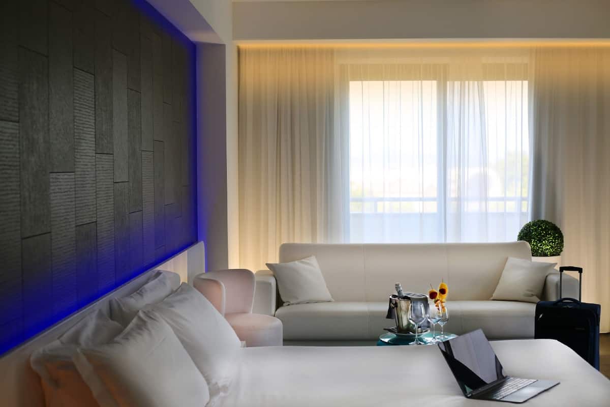 Une chambre d’hôtel avec un lit, un canapé, une petite table avec un ordinateur portable, une valise et une fenêtre avec des rideaux.