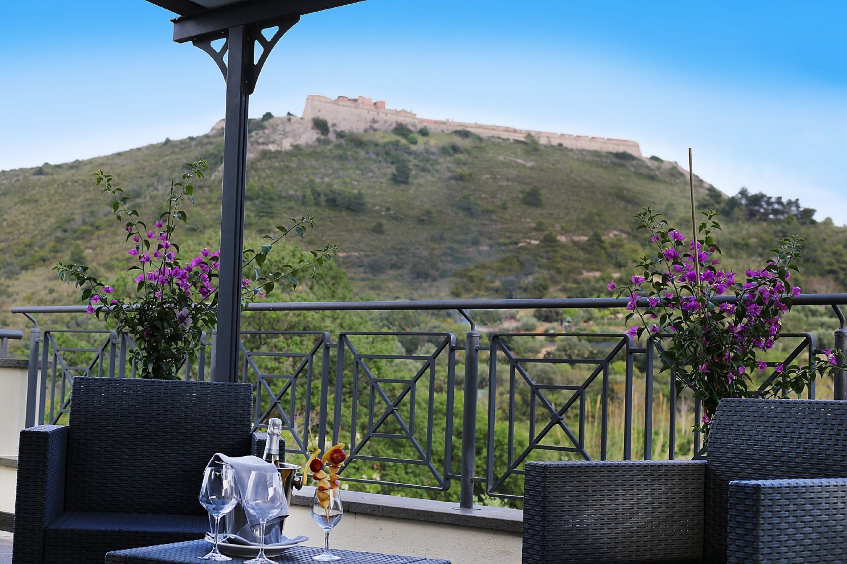 Una terraza con muebles de mimbre, una mesa con copas de vino y una bandeja de frutas, y vistas a una colina con una fortaleza.