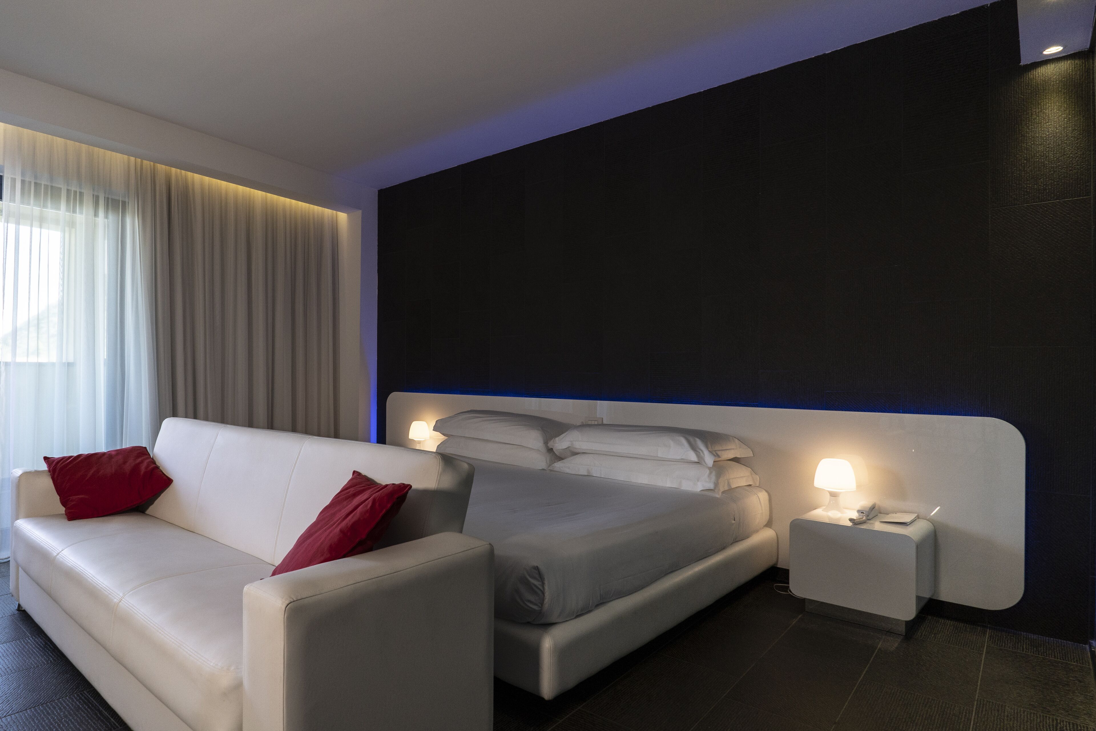 A modern hotel room with a large bed, a sofa, and a nightstand with a lamp.