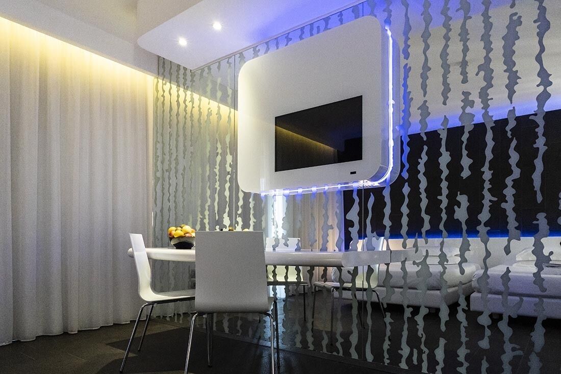 Quarto moderno com televisão de tela plana na parede, mesa de vidro com cadeiras e cortina estampada.