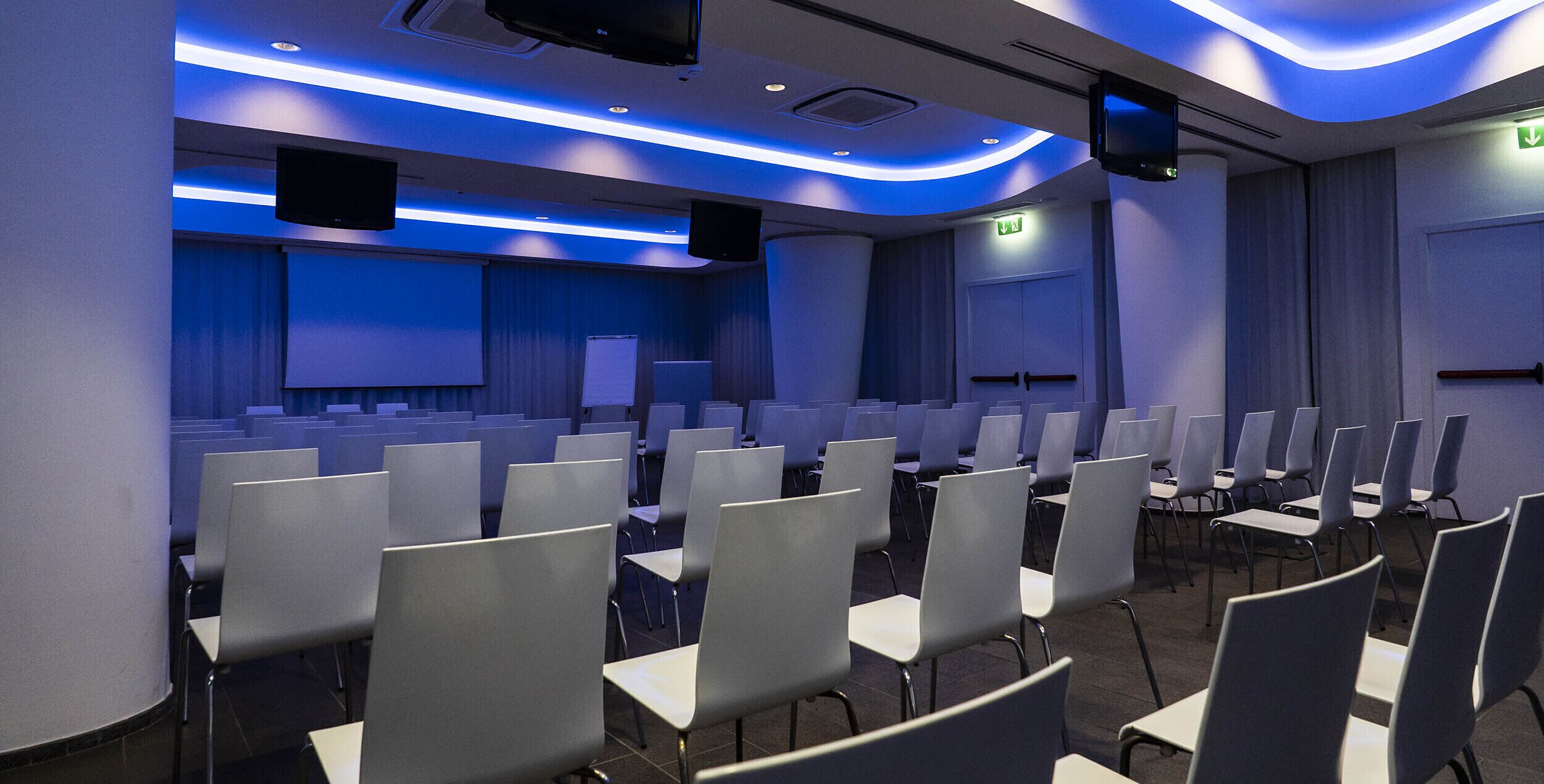Una sala de conferències amb files de cadires, una pantalla de projector i un escenari.