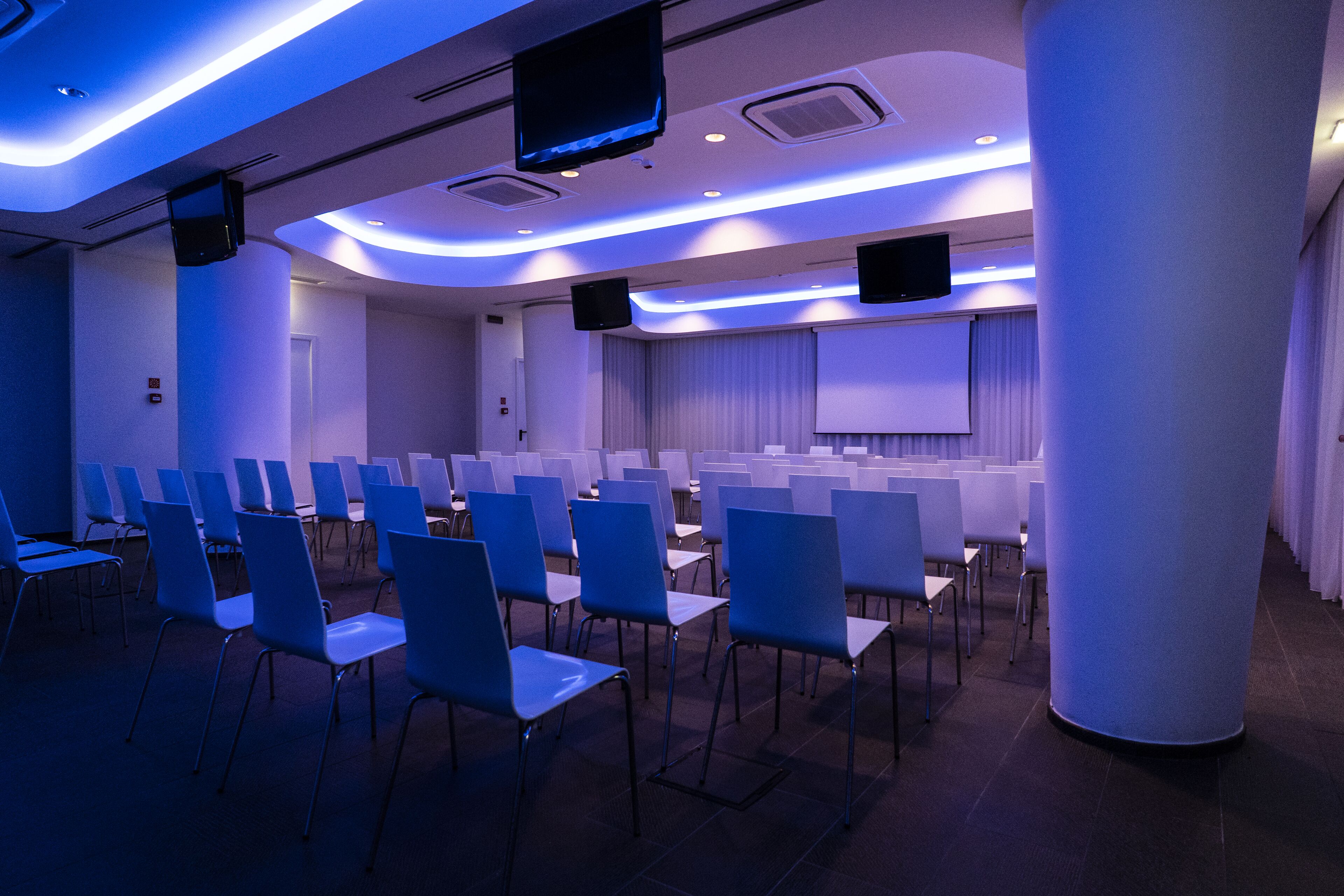 A conference room with rows of chairs, a large screen, and speakers on the ceiling.