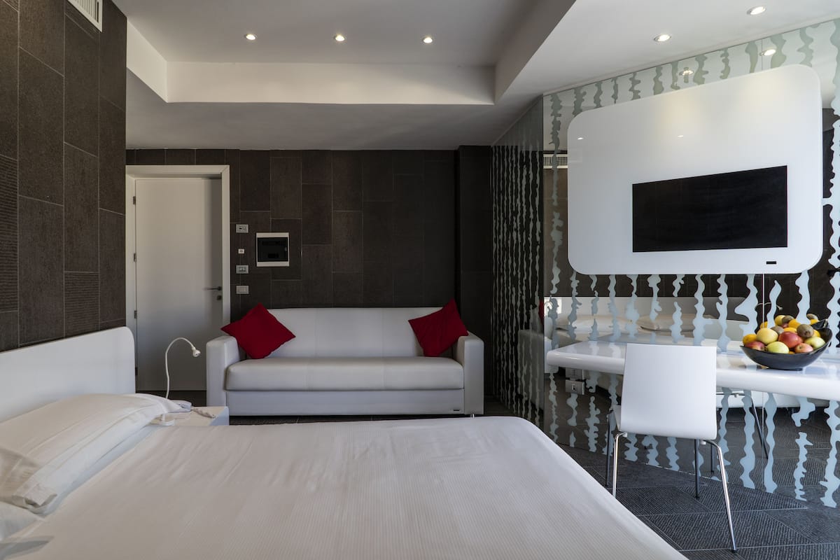 Une chambre d’hôtel moderne avec un grand lit, un canapé blanc agrémenté de coussins rouges, une table à manger avec un panier de fruits et un téléviseur fixé au mur.
