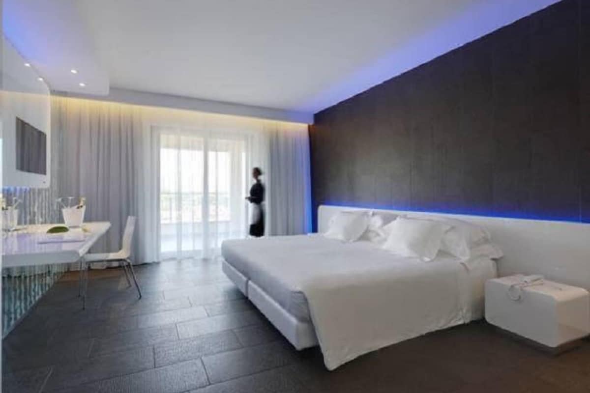 A modern hotel room with a large bed, a desk with a chair, and a large window with curtains.