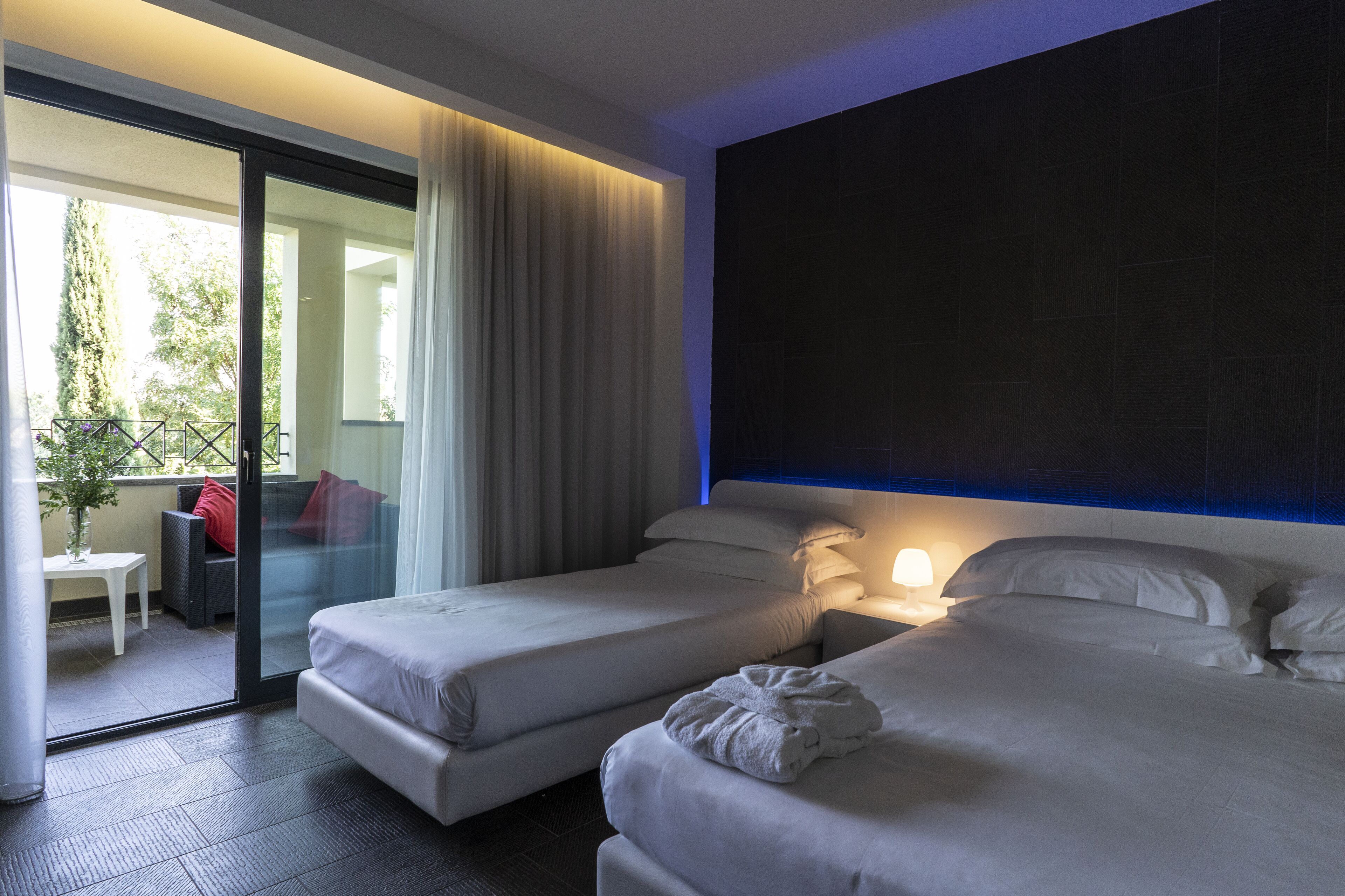 A hotel room with two beds, a sliding glass door to a balcony, a small table with a chair, and a nightstand with a lamp.
