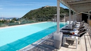 Una piscina azul cristalina con sillas de descanso y sombrillas a la derecha, rodeada por una terraza de madera y una colina al fondo.