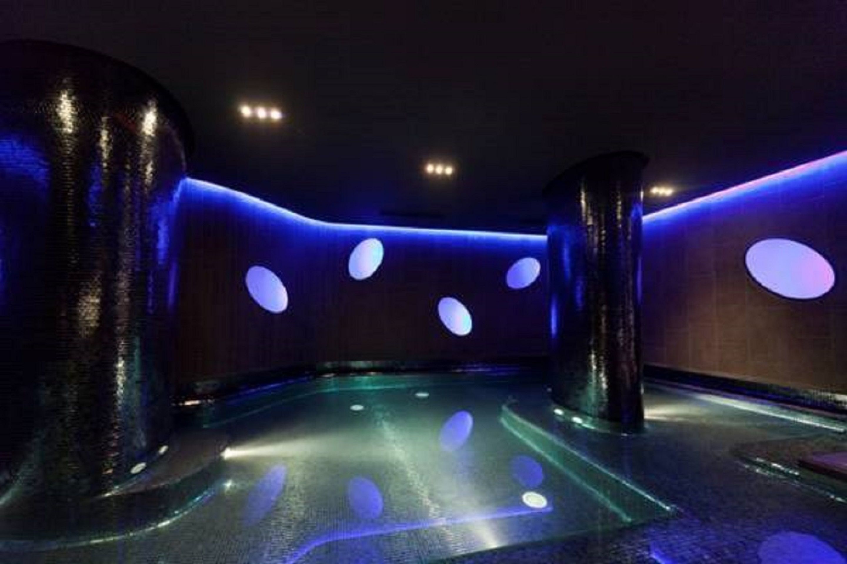 A spa room with a curved layout, illuminated by blue ambient lighting and featuring circular light fixtures on the walls.