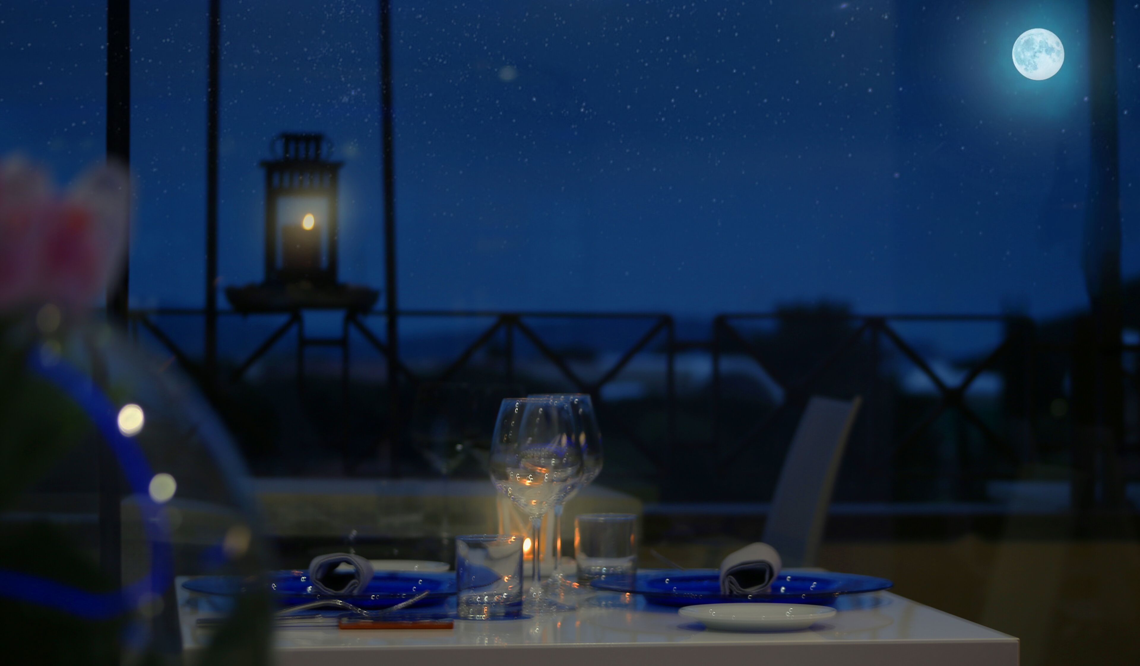 A table set for two with glasses and a candle, overlooking a night sky with a full moon and a distant lighthouse.