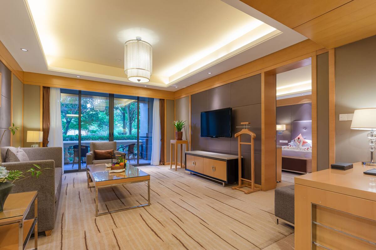 A modern living room with a glass coffee table, sofas, a TV, and a balcony with a view of greenery.