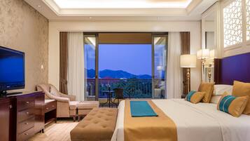 A hotel room with a large bed, a TV, a desk, and a balcony with a view of mountains.