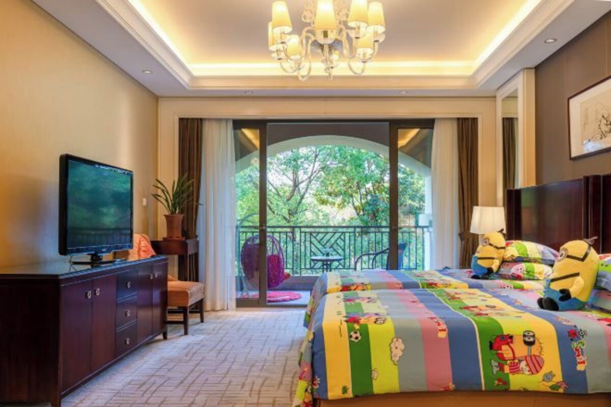 Une chambre d’hôtel avec un grand lit, une télévision, un balcon donnant sur la verdure et un lustre.
