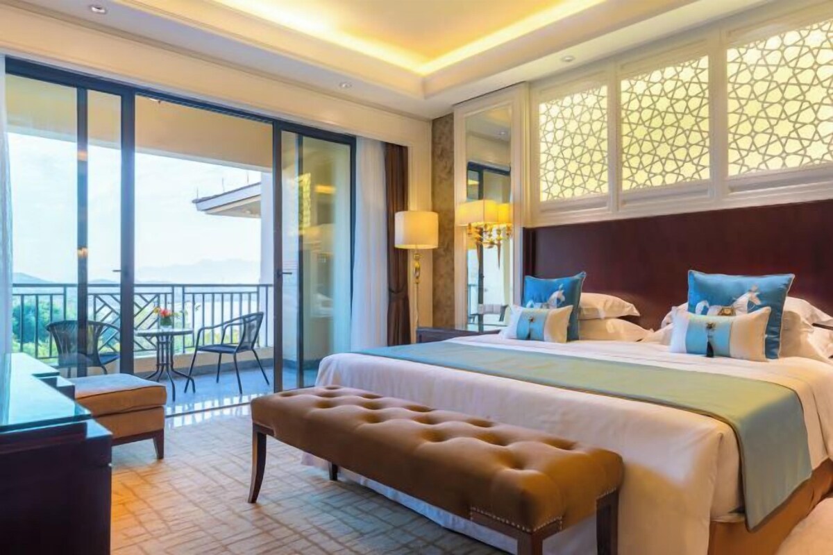 A hotel room with a large bed, a bench, a desk, and a balcony with chairs.