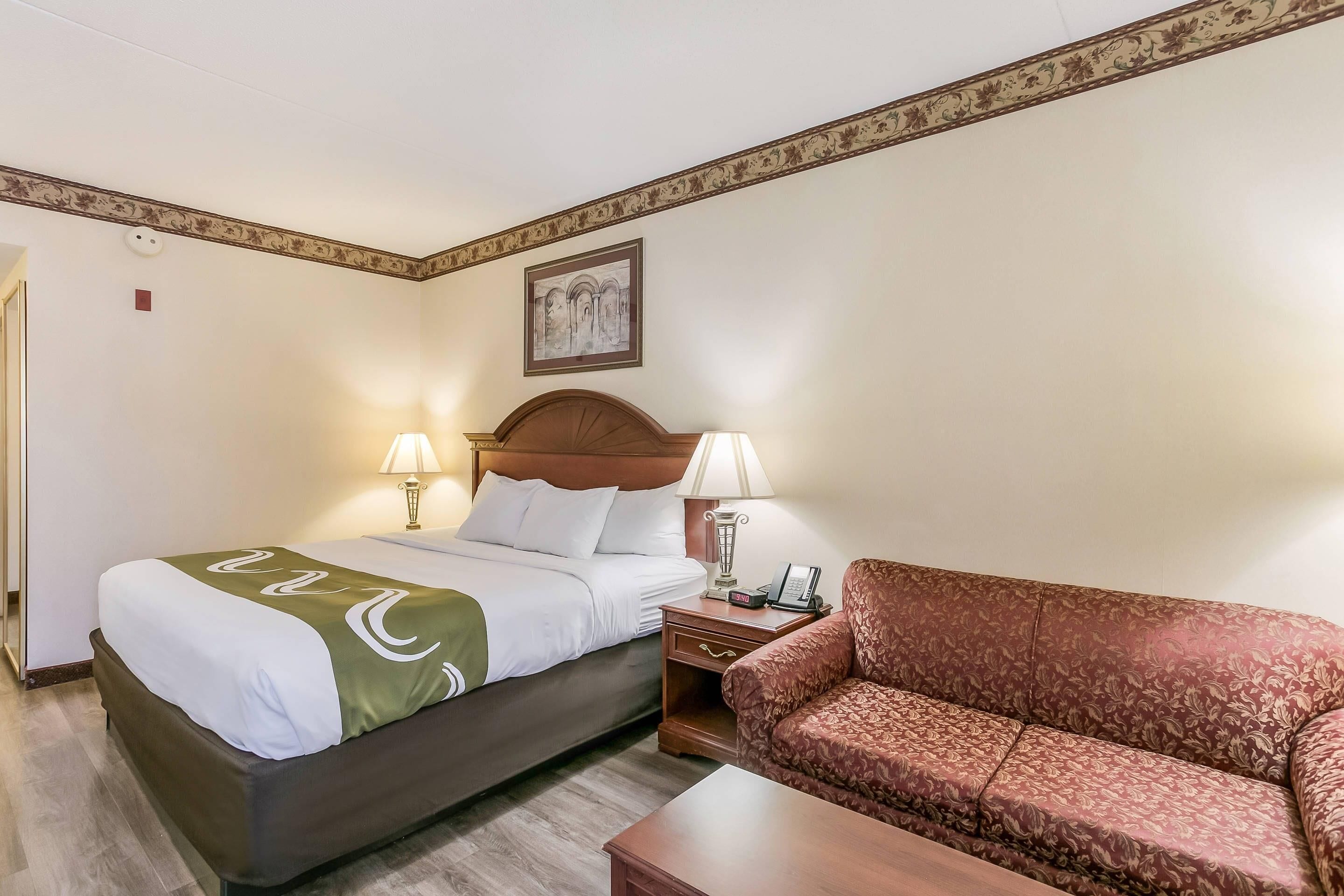 Une chambre d’hôtel avec un lit, deux lampes de chevet, un canapé, une table de chevet en bois et un tableau encadré au mur.