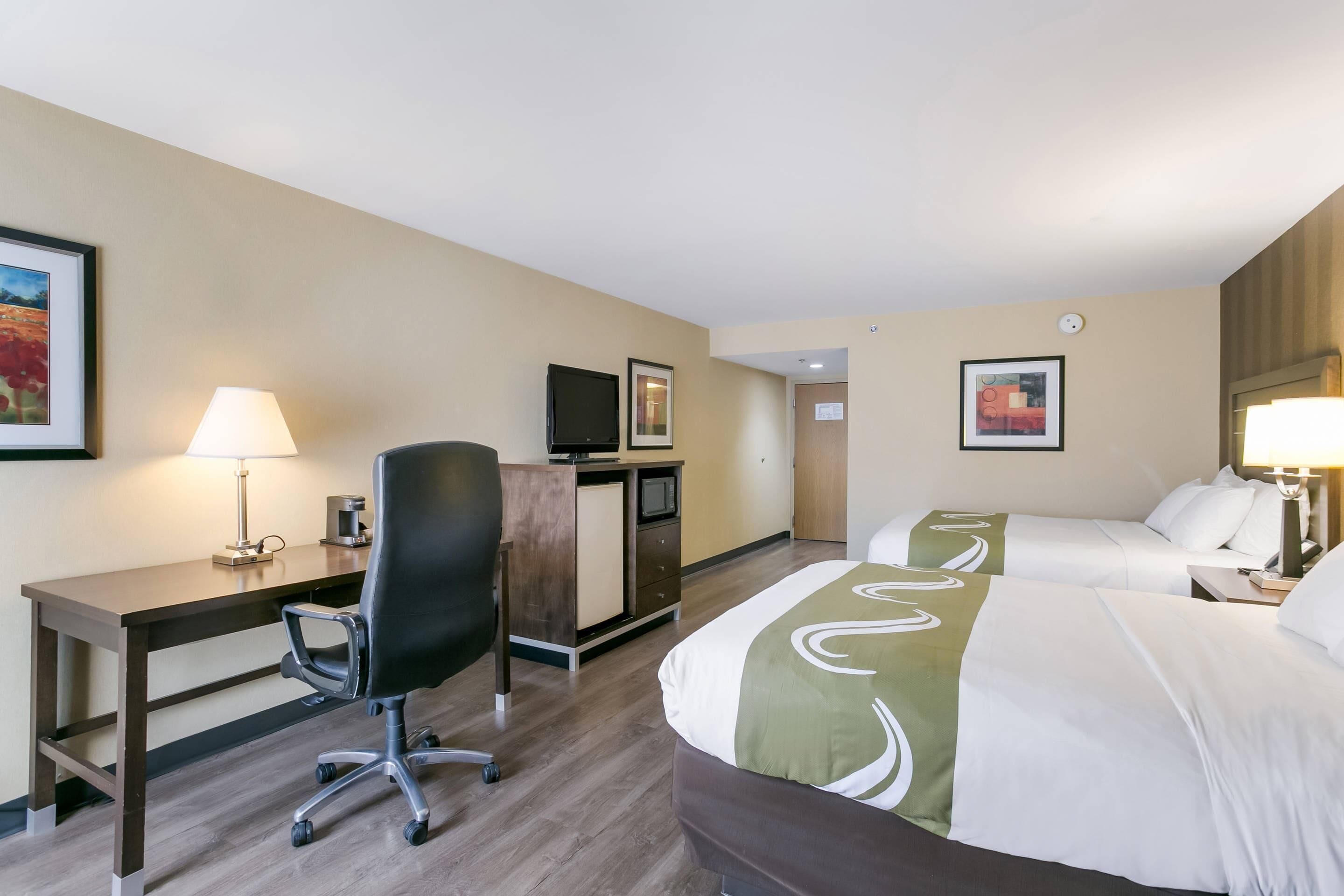 A hotel room with a desk, two beds, a chair, a TV, and artwork on the walls.