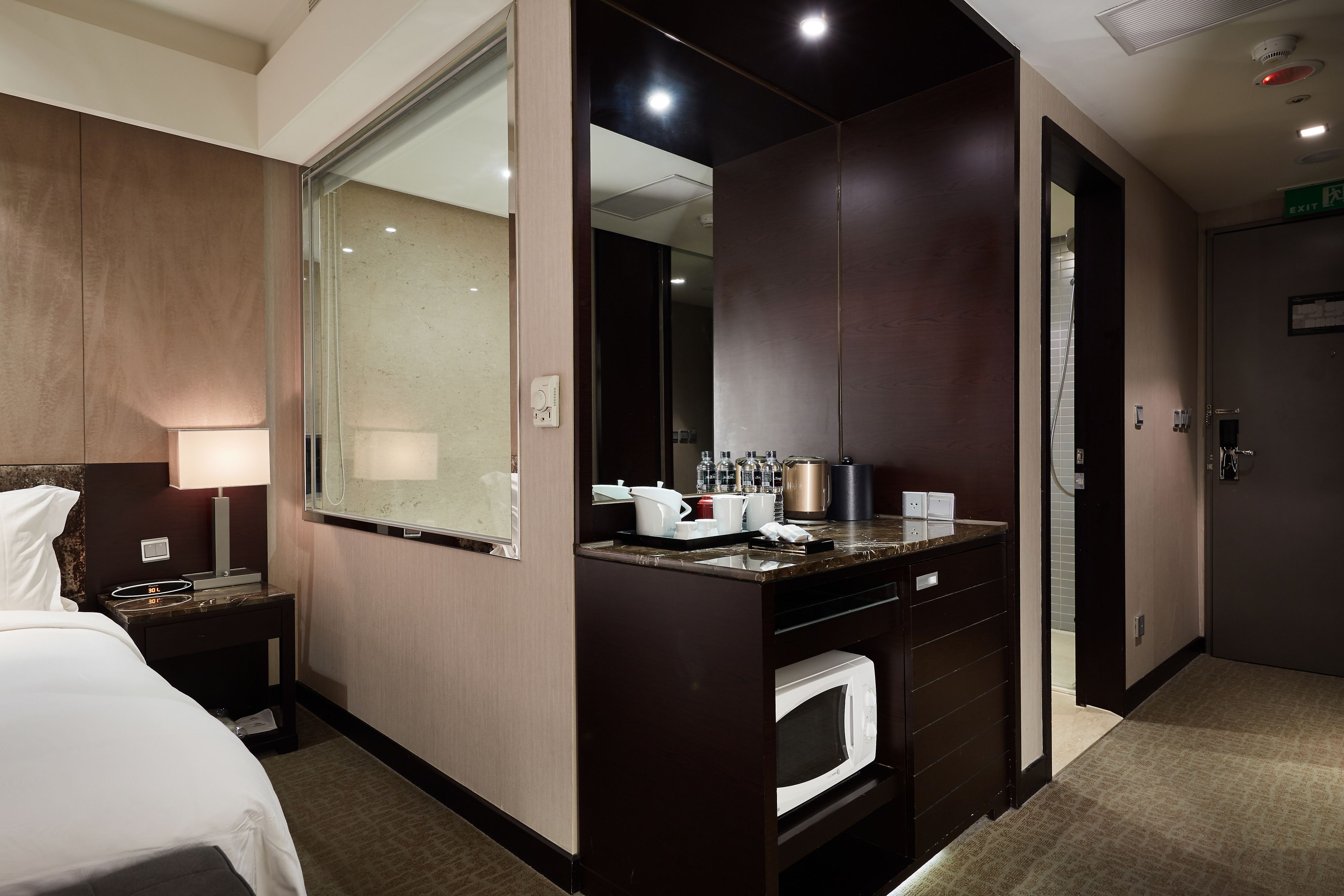 A hotel room with a bed, a desk with a lamp, a minibar, a microwave, and a large mirror.