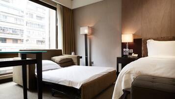 A hotel room with two beds, a desk, a lamp, and a view of a building through the window.