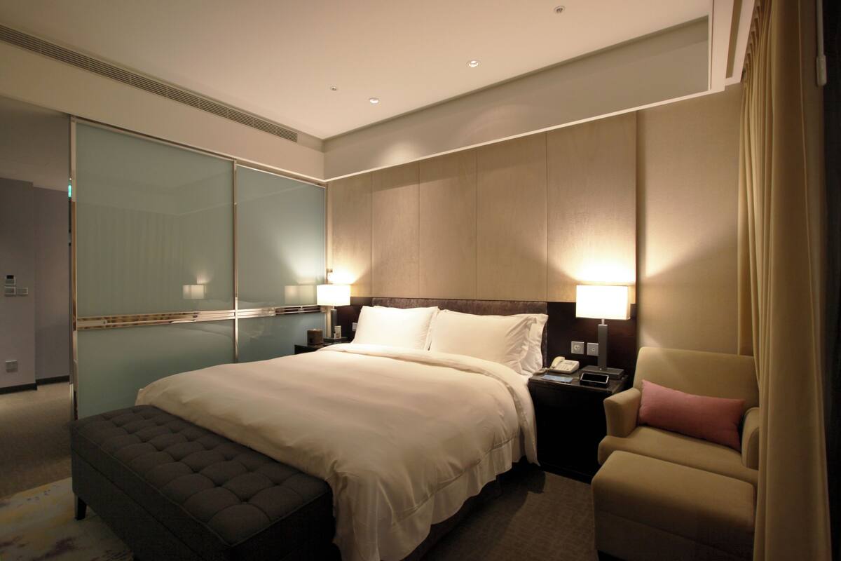 A hotel room with a large bed, a bedside table with a lamp, a chair, and a wardrobe.