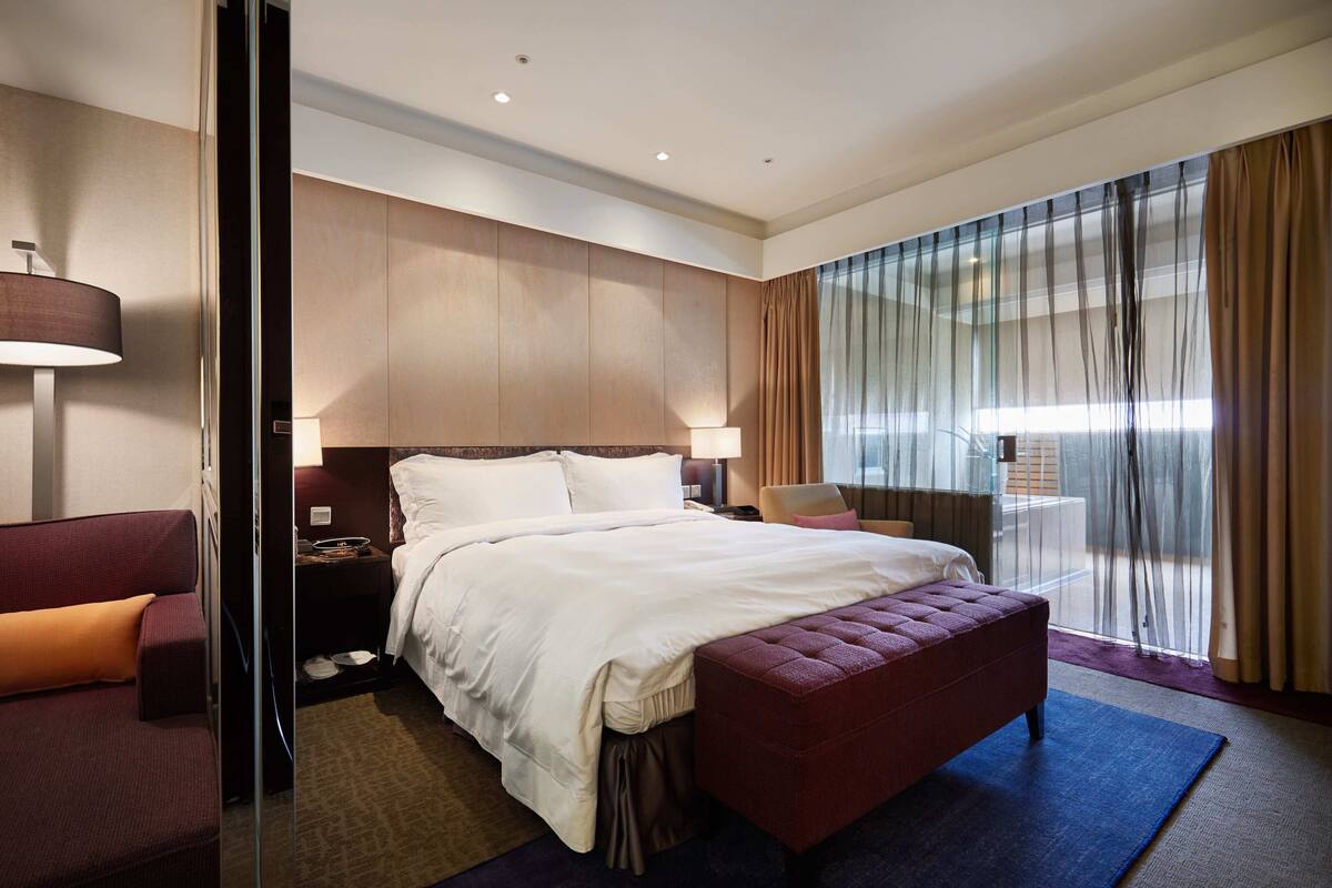 A modern hotel room with a large bed, a seating area with a red ottoman, a desk with a lamp, and a view of the outdoors through sheer curtains.