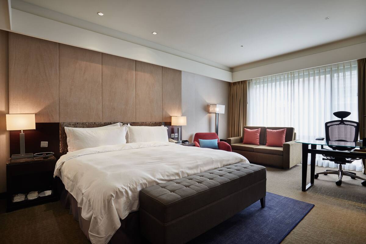 A hotel room with a large bed, a desk with a chair, a sofa, and a window with curtains.