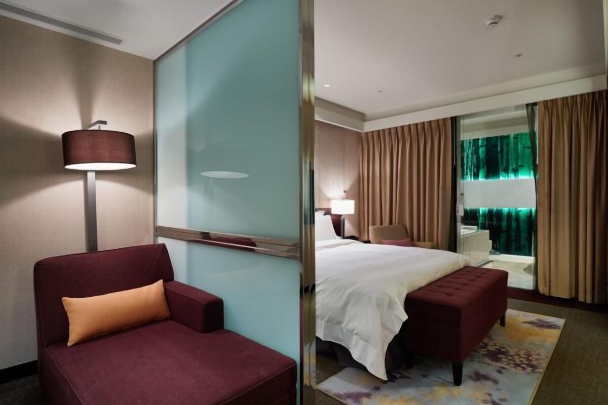 A hotel room with a bed, a red armchair, a floor lamp, and a large window with curtains.