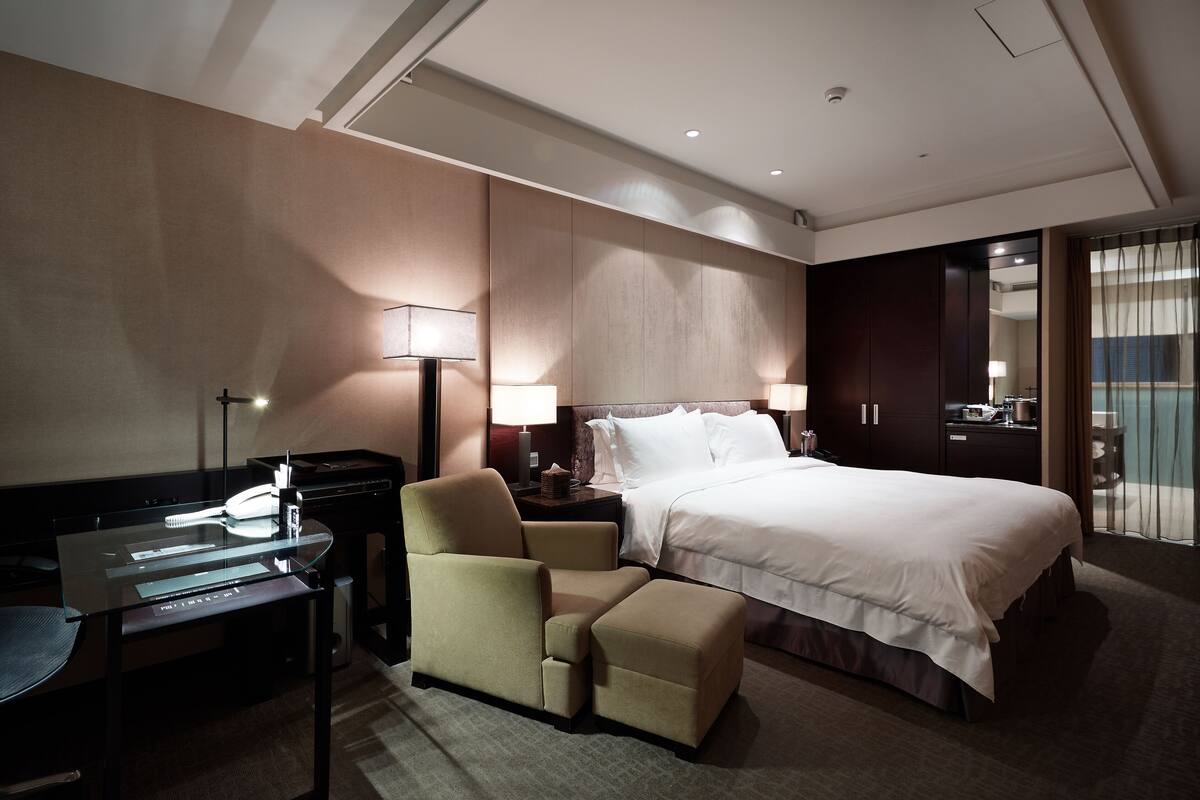 A hotel room with a bed, a desk, a chair, and a nightstand with lamps.