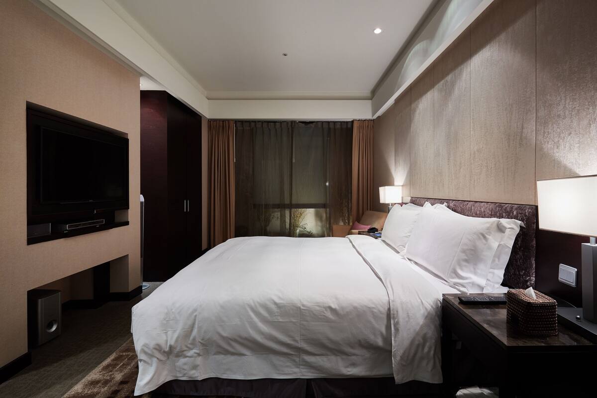 A hotel room with a large bed, a flat-screen TV, a bedside table with a lamp, and a window with curtains.