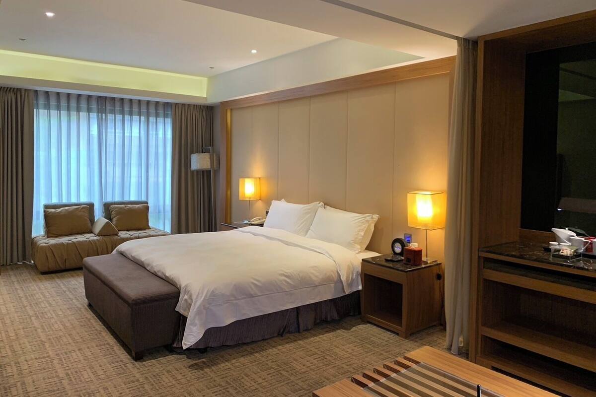 A hotel room with a large bed, two bedside lamps, a TV, a sofa, and a desk with a lamp.