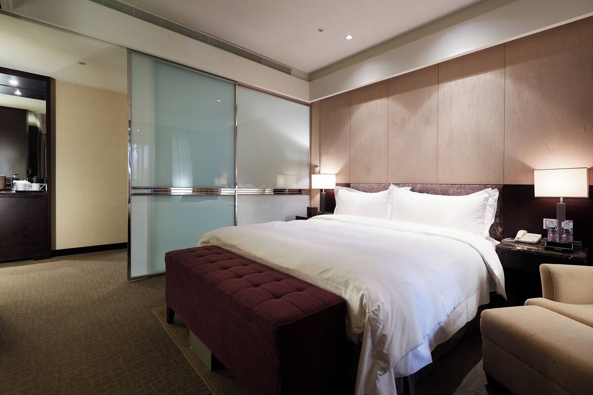 A hotel room with a large bed, a bedside table with a lamp, a chair, a small table with a phone and a bottle, and a wardrobe with a mirror.